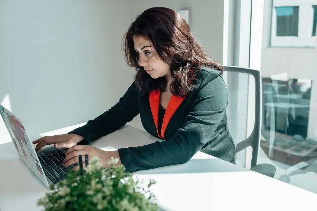 A woman in a dark blazer typing on a laptop at a bright office desk