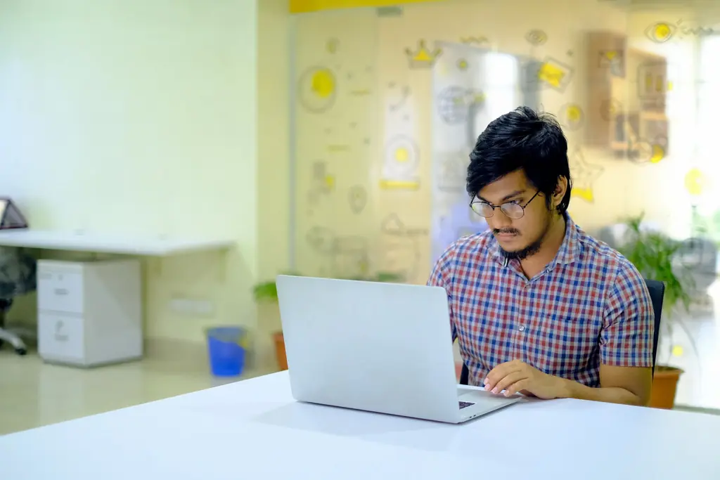 A man in glasses and a checked shirt working on a laptop in a bright, open office